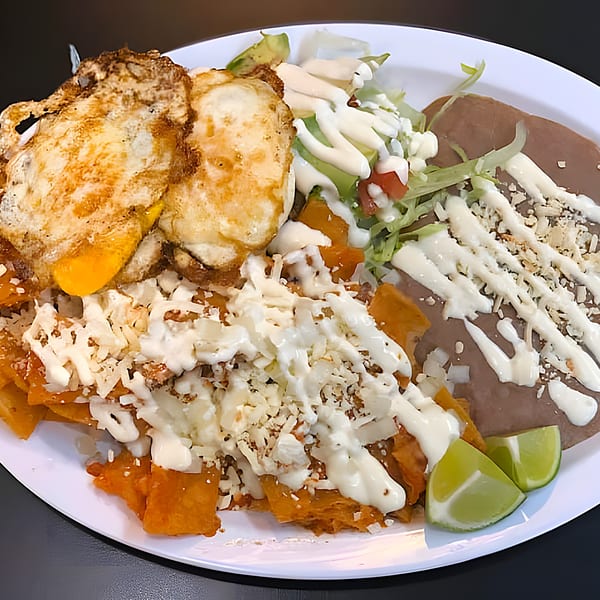 Chilaquiles with fried eggs, refried beans, lettuce, avocado, and lime wedges on a white plate.