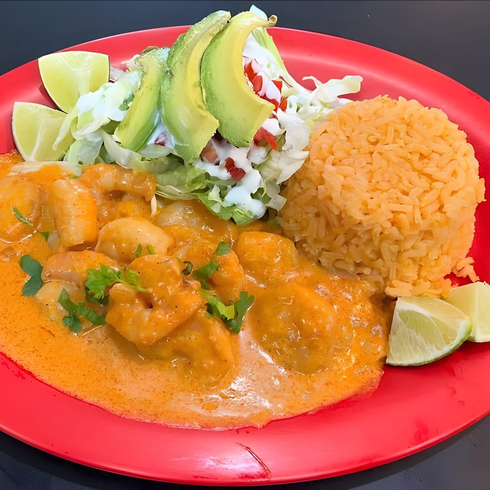 Shrimp in orange sauce with rice, avocado, lettuce, and lime wedges on a red plate.