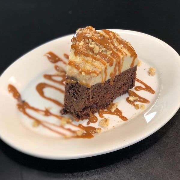 A slice of chocolate cake with a light-colored topping, drizzled with caramel and sprinkled with nuts.