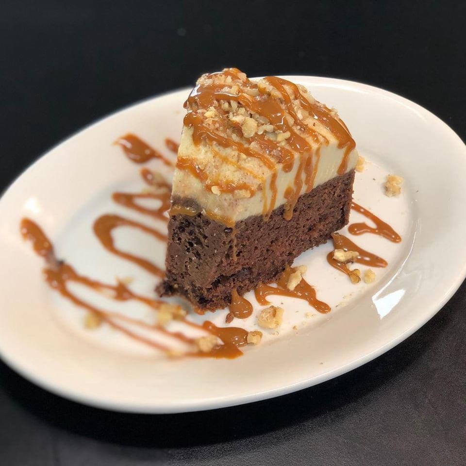 A slice of chocolate cake with a light-colored topping, drizzled with caramel and sprinkled with nuts.