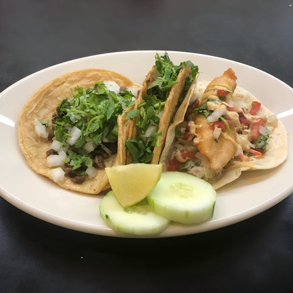 Three Nacho's Taqueria Grill tacos, two with meat and one fish, garnished with lime and cucumber.