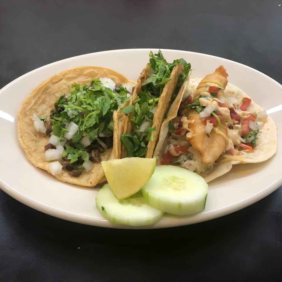 Three Nacho's Taqueria Grill tacos, two with meat and one fish, garnished with lime and cucumber.